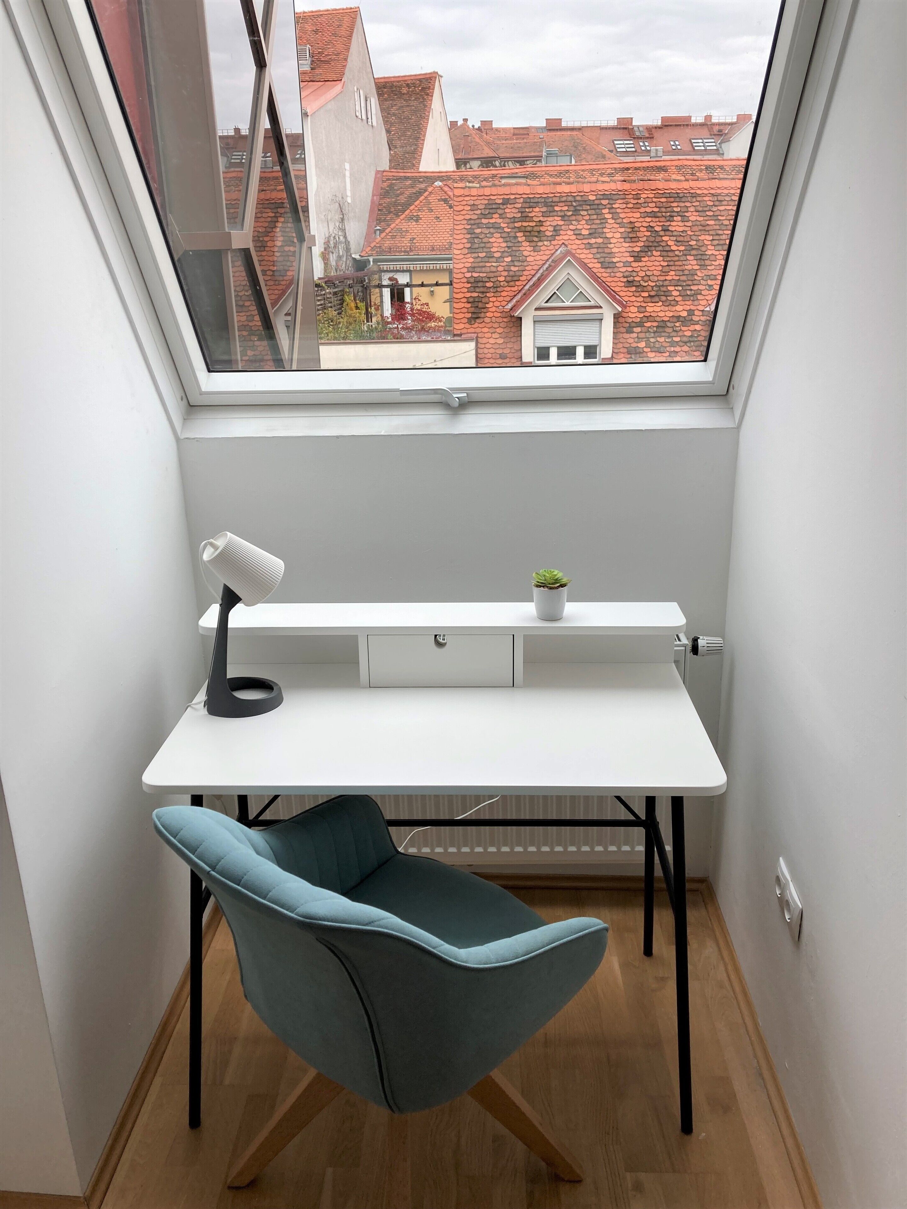 Une pièce d’angle avec un puits de lumière, un bureau blanc, une chaise de couleur turquoise et une petite plante.