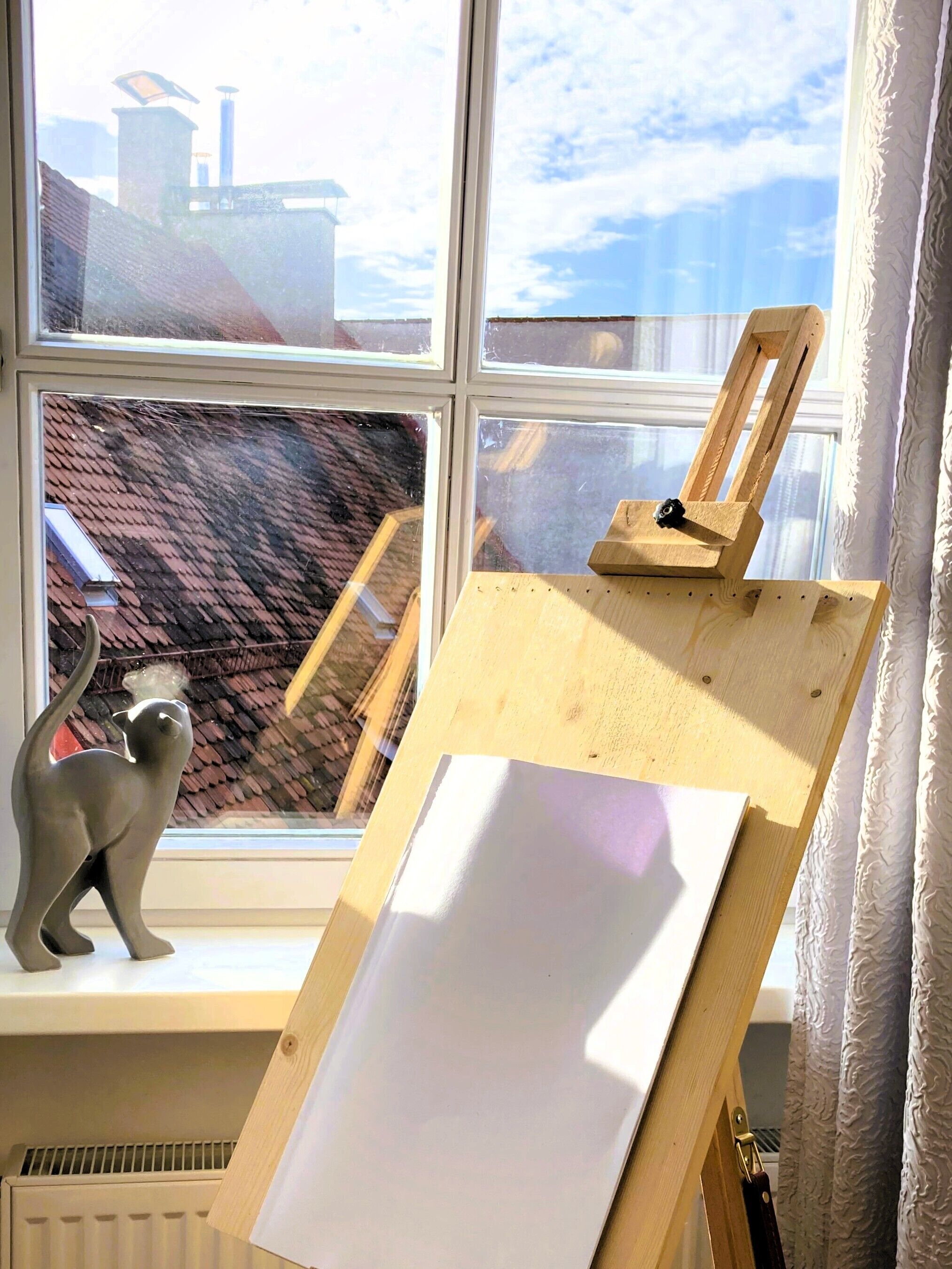 Um quarto com uma janela, um cavalete de madeira com tela em branco, e uma estatueta de gato no parapeito.