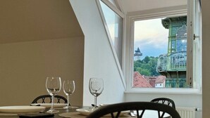 A dining area with a table set for two, a window with a view of a green building, and a sloped ceiling. - Carillon by BestChoice  - Parking Option (Graz)