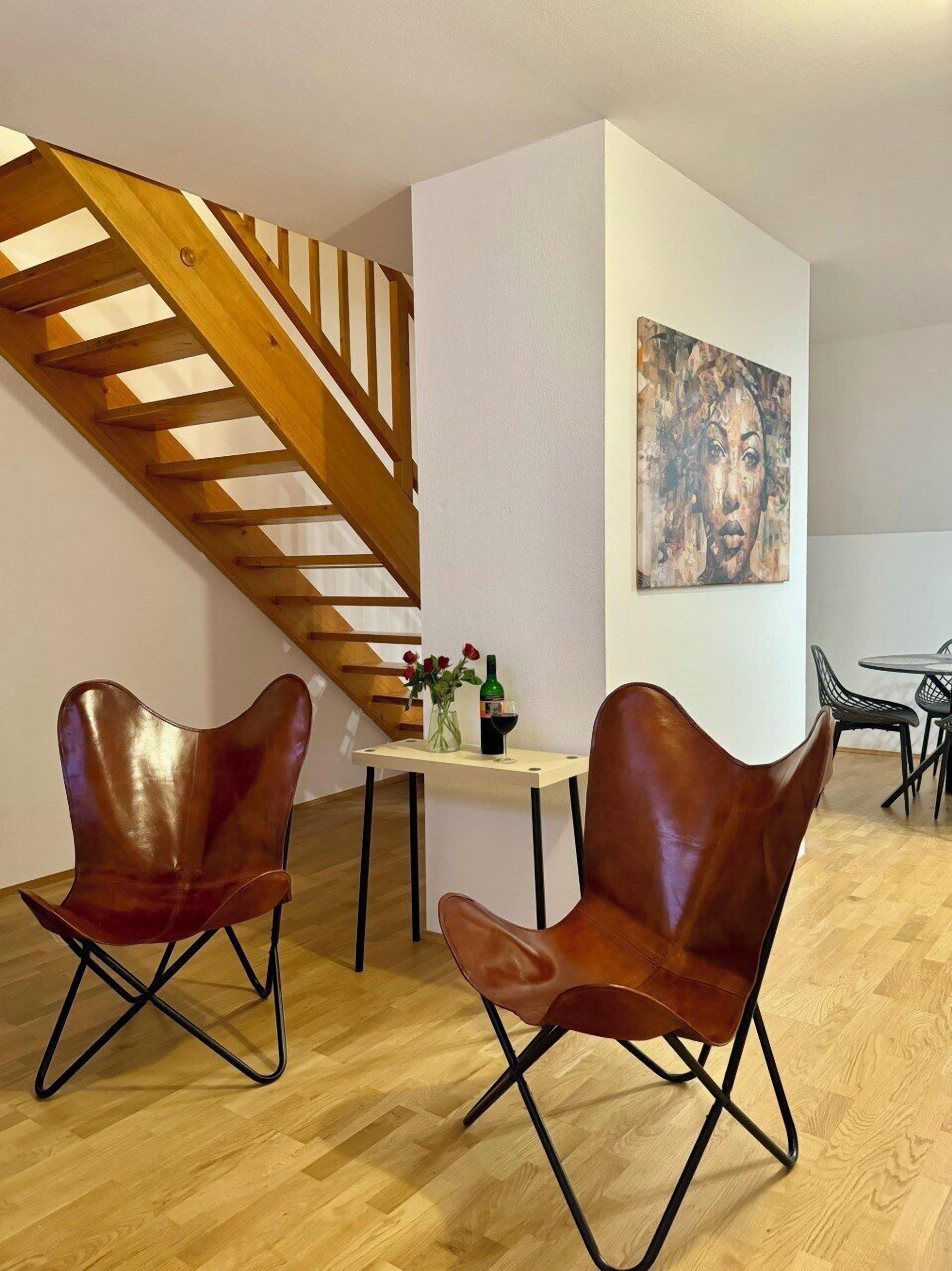 Un salon moderne avec un escalier en bois, deux fauteuils en cuir, une petite table sur laquelle se trouvent un vase et une bouteille de vin, et un tableau accroché au mur.