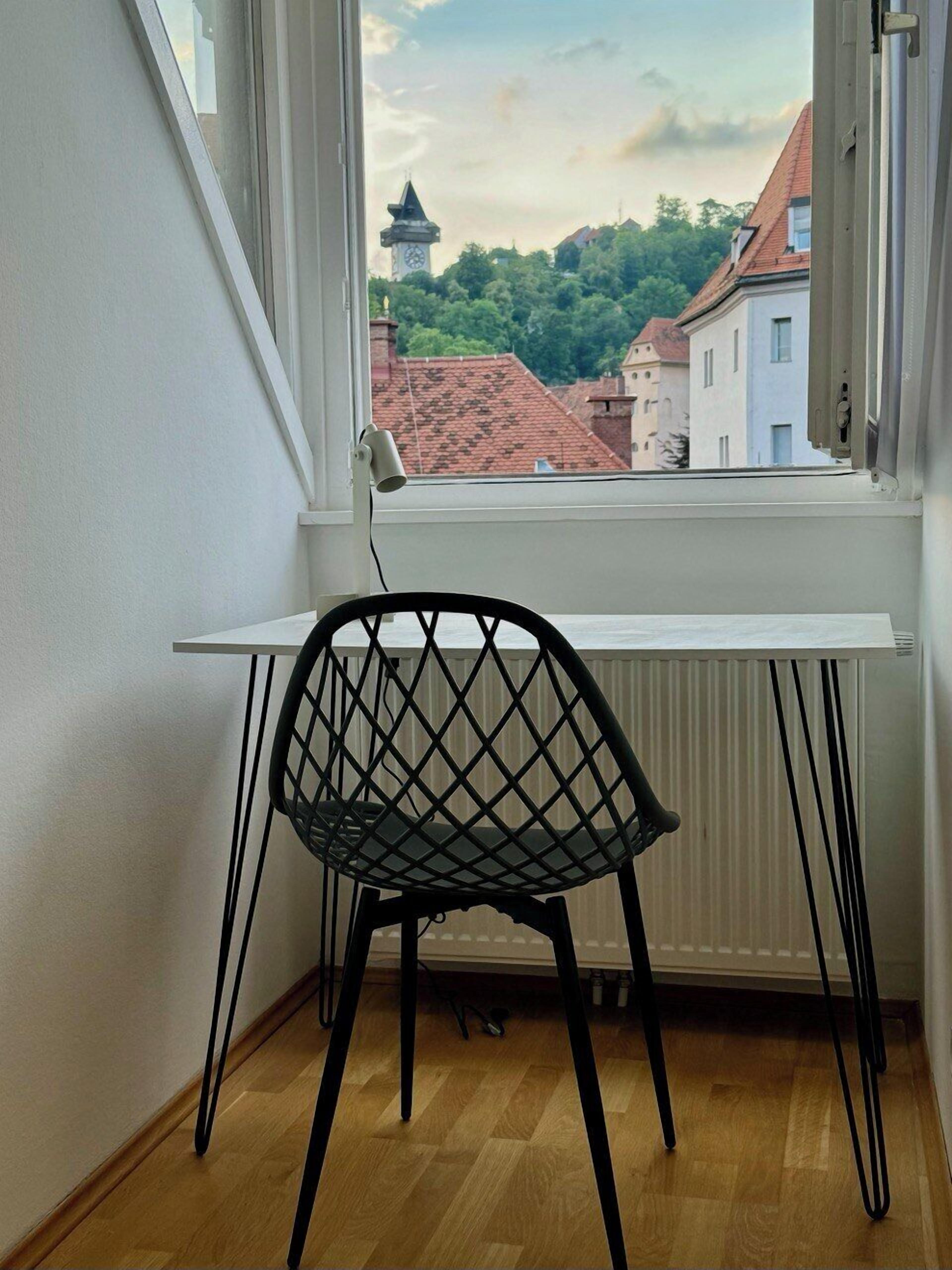 A corner room with a white desk, a black chair, and a view of a town with buildings and a tower.