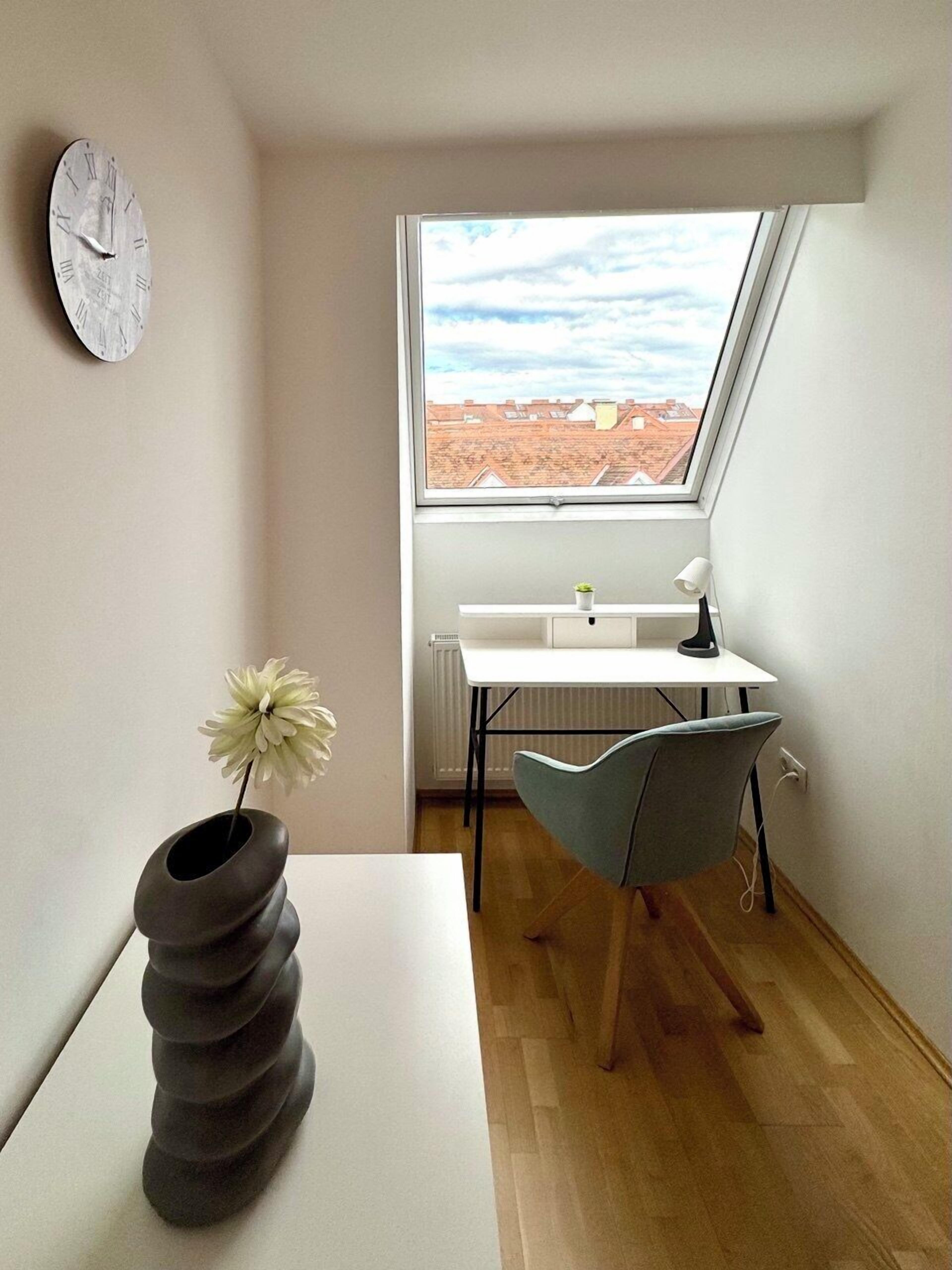 A modern room with a sloped ceiling, a skylight, a white desk, a grey chair, a white radiator, and a wall clock.