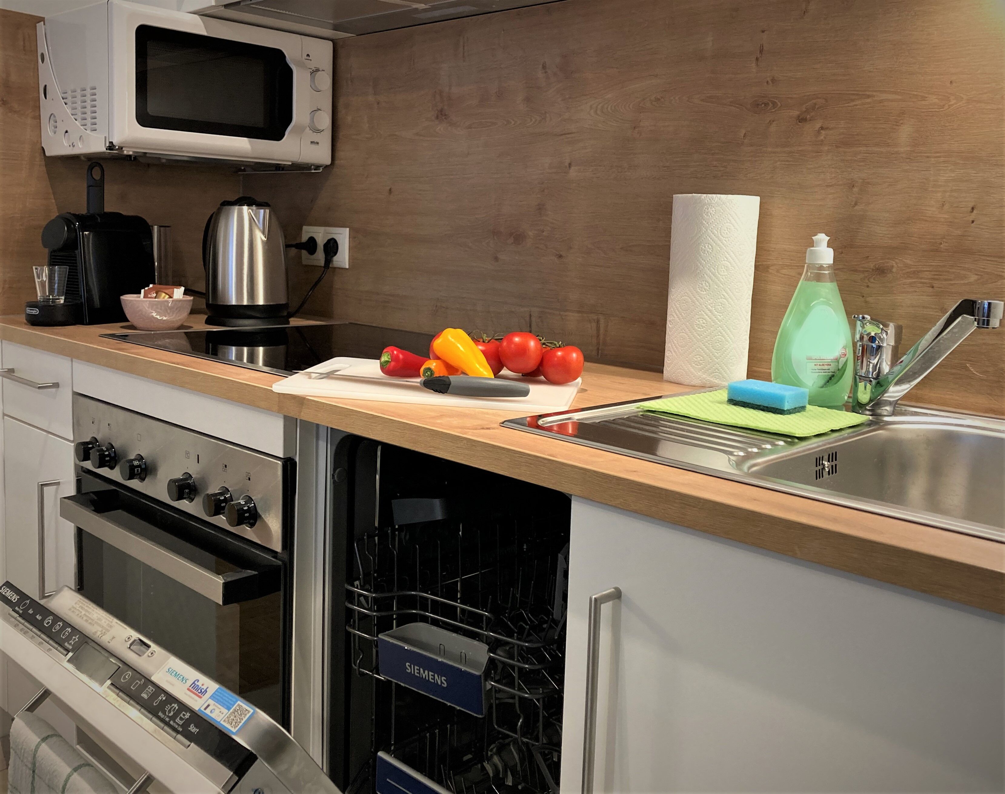 A modern kitchen with a microwave, kettle, and a cutting board with vegetables.