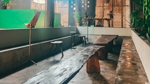 A spacious indoor area with a long wooden bench, a small table, and a seating area with stools. There are potted plants and a bamboo fence visible. - Varuna Home (Tulum)