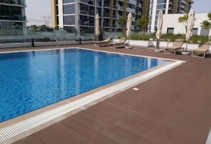 A rectangular swimming pool with a clear blue water surface, surrounded by a tiled deck area with lounge chairs and umbrellas. - Bloomfields Lovely 1BHK Azizi Riviera (Dubai)