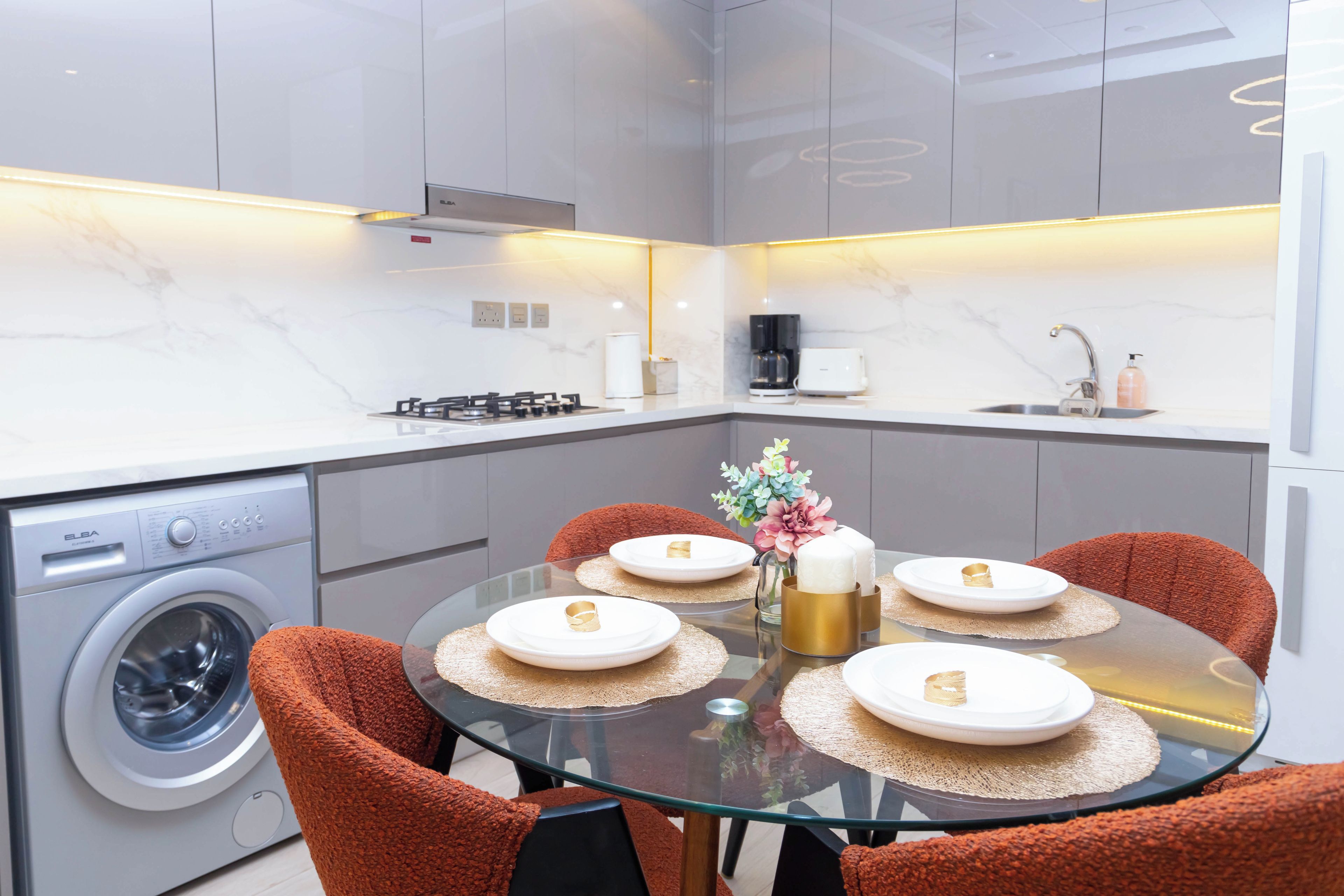 Une cuisine moderne équipée d’un lave-linge intégré, d’une machine à café et d’un coin repas avec une table en verre et des chaises orange.