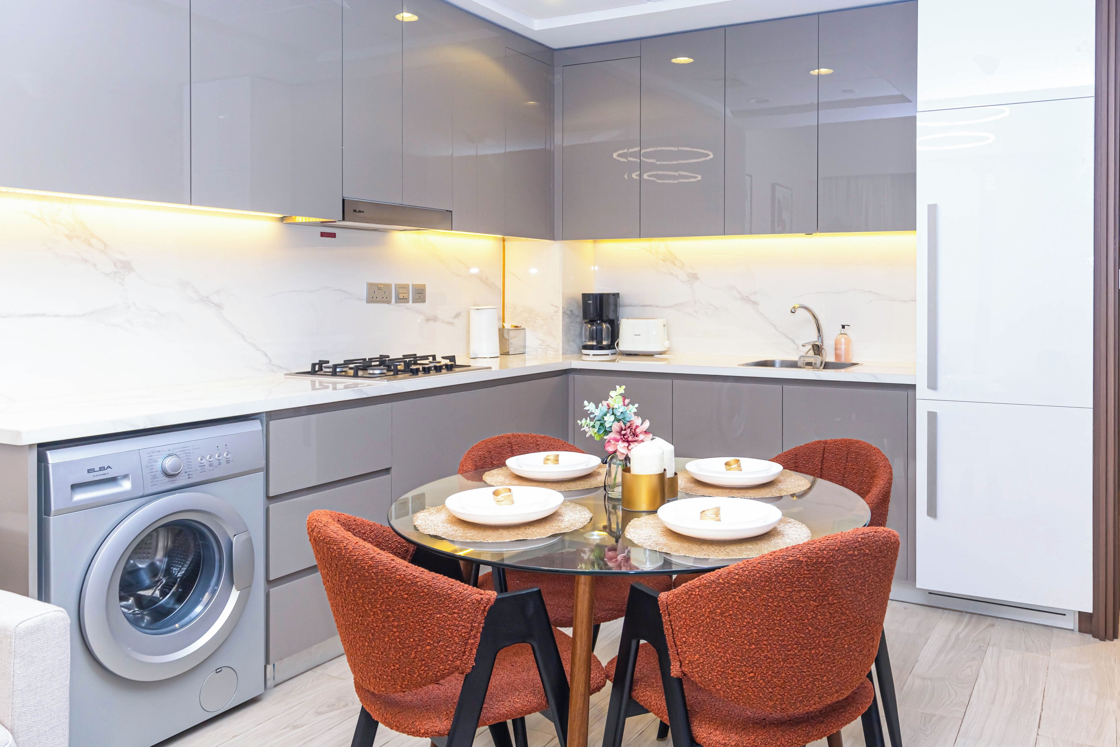 A modern kitchen with a built-in washing machine, a dining area with a glass table and orange chairs, and a refrigerator.