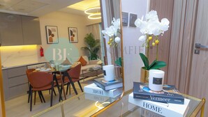 A modern hotel room with a glass table, a sofa, and a dining area with chairs. There are books and a candle on the table.