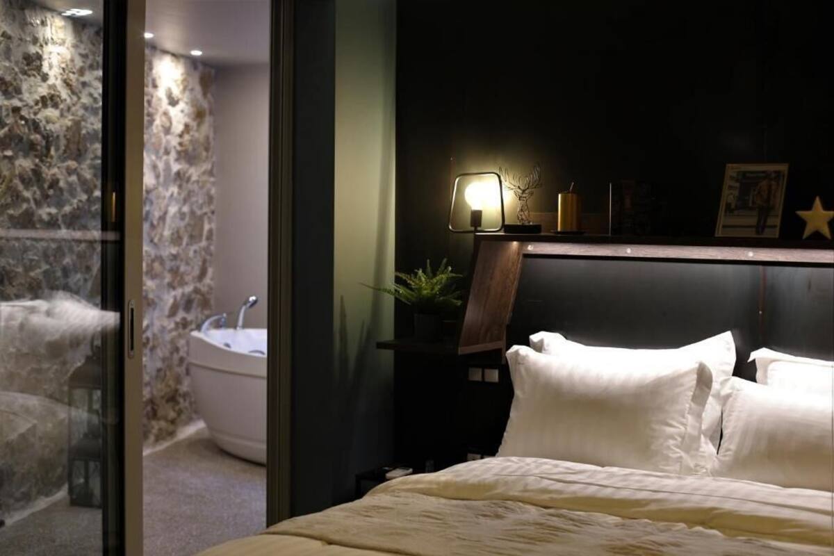 A hotel room with a bed, a nightstand with a lamp, a wall-mounted shelf with decorative items, and a bathroom with a bathtub and a sink.