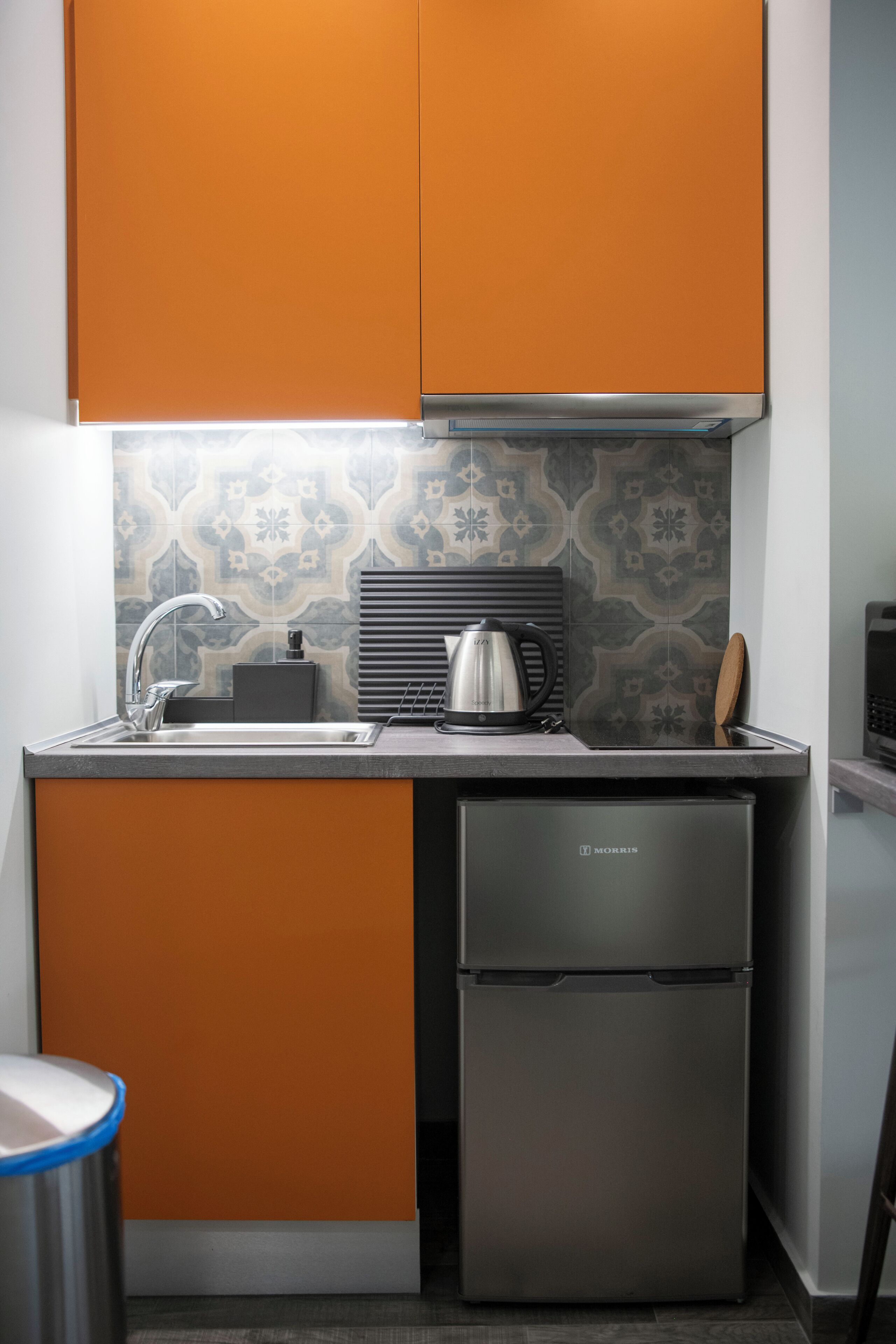 A compact kitchen with orange cabinetry, a stainless steel refrigerator, and a patterned backsplash.
