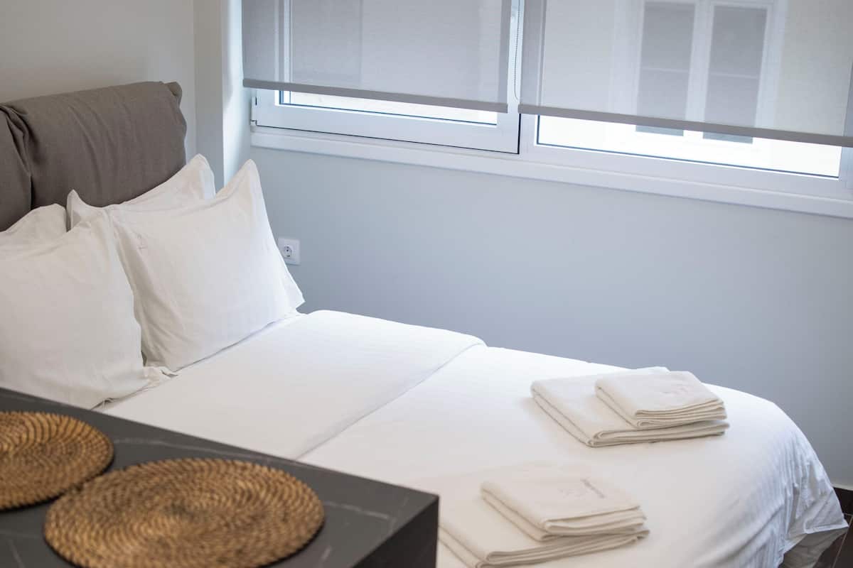 A neatly made bed with white linens, a small table with a woven placemat, and a window with blinds partially open.