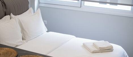 Un lit bien fait, avec du linge de lit blanc, une petite table sur laquelle repose un set de table en fibres tressées, et une fenêtre dont les stores sont entrouverts.