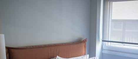 A neatly made bed with white linens, a wooden nightstand with woven placemats, a black radio, and a window with blinds.