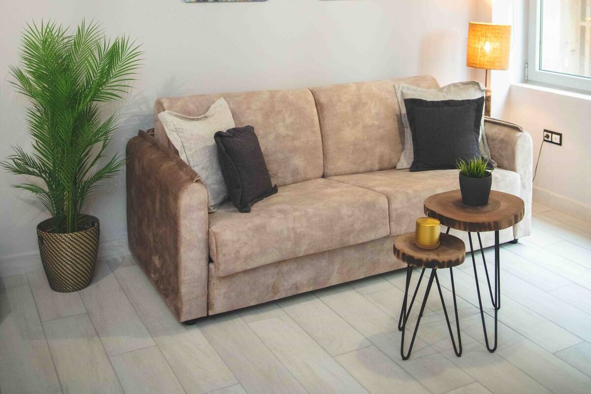 A living room with a beige sofa, two wall-mounted paintings, a potted plant, a small round table with a lamp, and a window with natural light.
