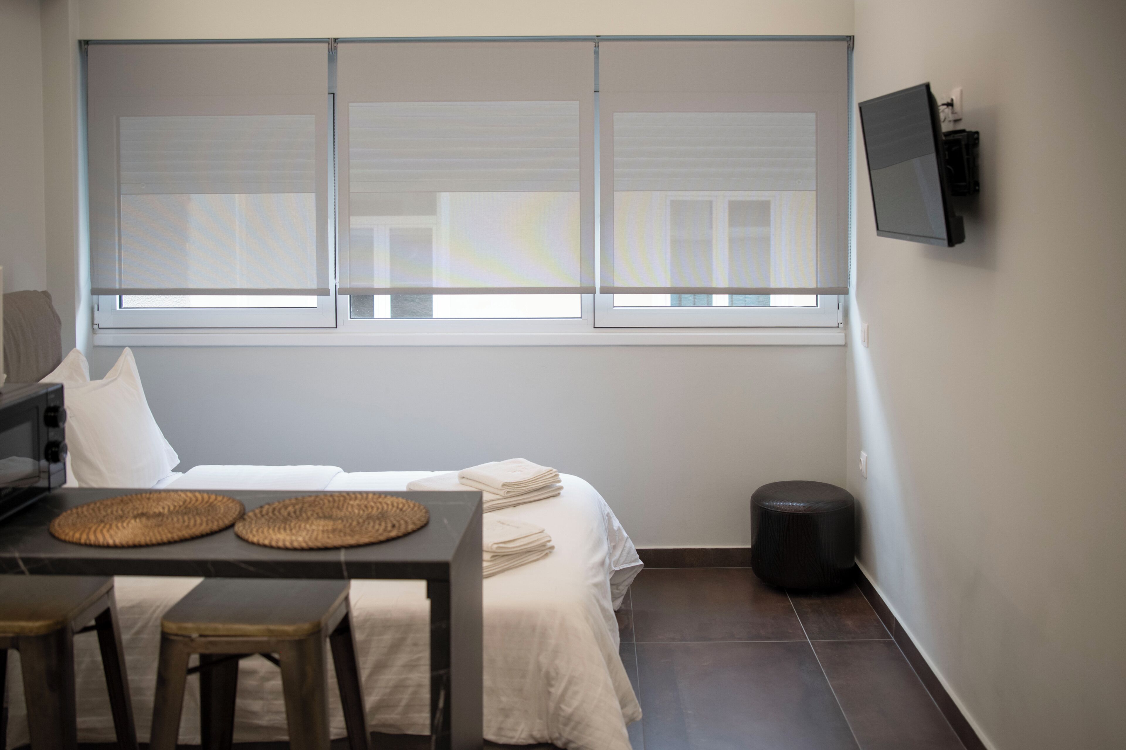 Ein Hotelzimmer mit einem Bett, einem Tisch mit Hocker, einem an der Wand befestigten Fernseher und einem Fenster mit Jalousien.