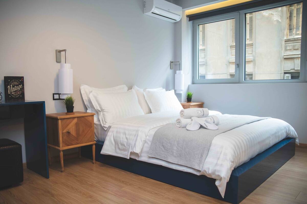 A modern bedroom with a bed, bedside tables, a wall-mounted lamp, and a window with a view of buildings.