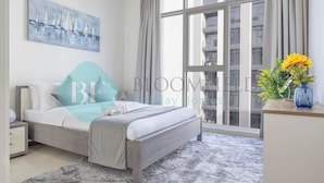 A modern bedroom with a large window, a bed with grey bedding, a bedside table with a lamp, and a painting on the wall. - Bloomfields Metropolitan 3BR In Bridges (Abu Dhabi)