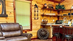 A cozy living room with a leather sofa, a round coffee table with a book, a rustic wooden floor, and a kitchen area with bar stools and a sink. - 𝐁 𝐢𝐠 𝐒𝐨𝐮𝐭𝐡 𝐅𝐨𝐫𝐤 Retreat! Excellent Location•Private•WIFI, 75” TV, Horses Welcome! (Strunk)