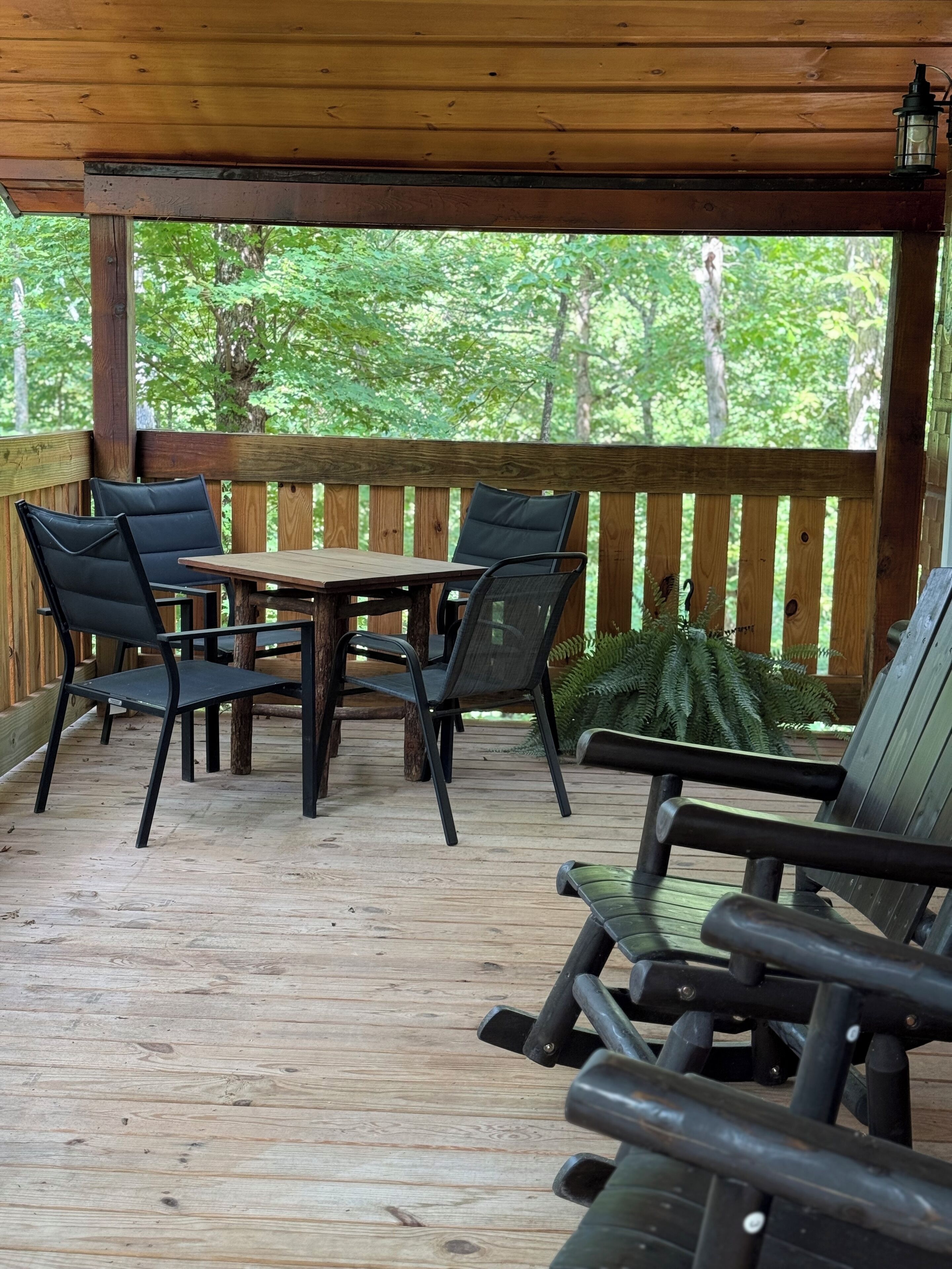 A wooden deck with a table and chairs, surrounded by a wooden railing, overlooking a forested area.