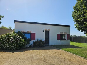 Un pequeño edificio de una sola planta con una puerta y dos ventanas, rodeado por un camino de grava y una zona ajardinada.