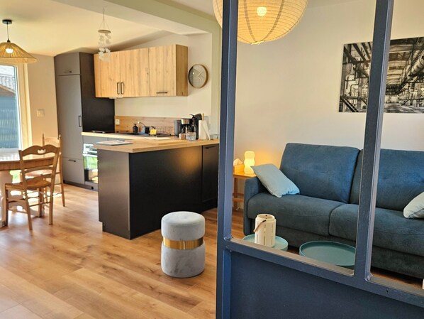 Una cocina moderna con armarios de madera, un comedor con una mesa y sillas de madera, un otomán gris y un sofá azul con cojines.
