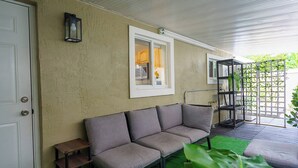 A covered patio area with a gray sofa, a small table, and a window with a view of the interior.