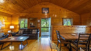 A cozy cabin interior with a wooden ceiling, a sofa, a dining table, and a view of the outdoors through a door. - Big Oak Cabin (Hocking Hills area) (Rockbridge)
