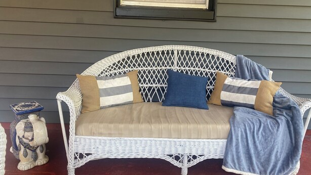 A wicker sofa with cushions, a small bear figurine, and a framed picture on a porch with gray siding.