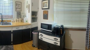 A compact kitchen with a sink, stove, and microwave. There is a trash bin and a small cabinet with drawers.