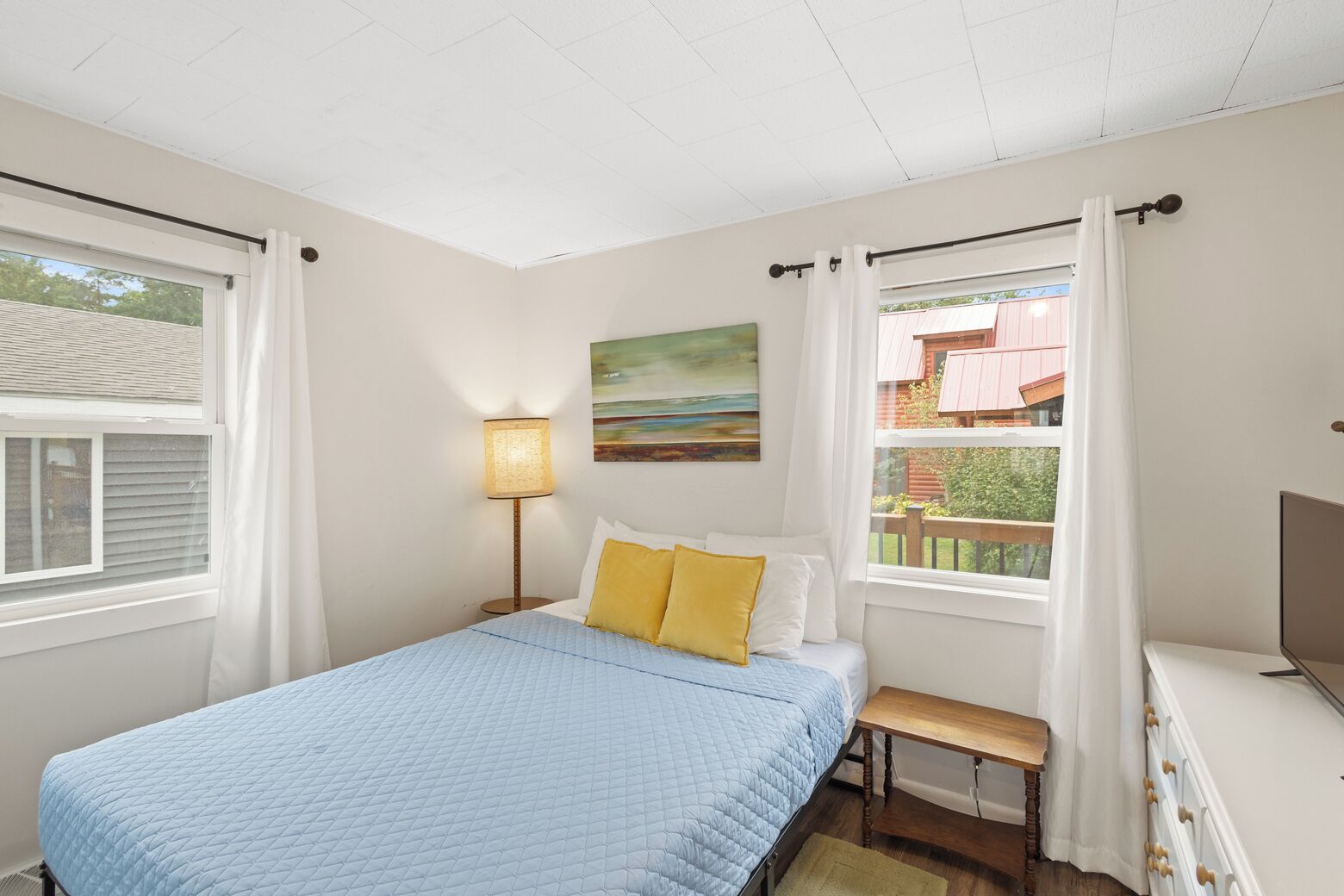 A bedroom with a bed, a bedside table, a lamp, a painting, and a window with curtains.