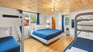 Habitación con literas, techo de madera, cama con manta azul, ventana con vistas a árboles y un escritorio pequeño con lámpara.