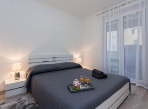 A modern bedroom with a large bed, bedside lamps, a tray with glasses and orange juice, and a view through sheer curtains. - Stylish Zadar Apartments With Easy Access to Old Town (Zadar)