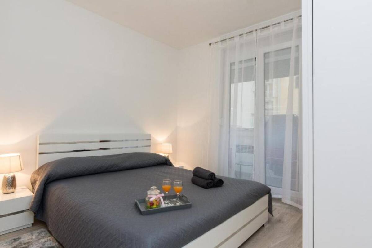 A modern hotel room with a large bed, a bedside table with a lamp, and a tray with drinks and a small bottle.
