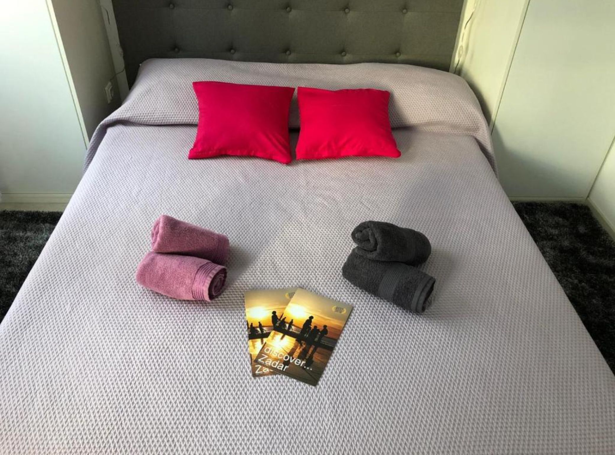 Een net opgemaakt bed met twee rode kussens, twee roze en twee donkergrijze handdoeken, en een brochure op het bed.