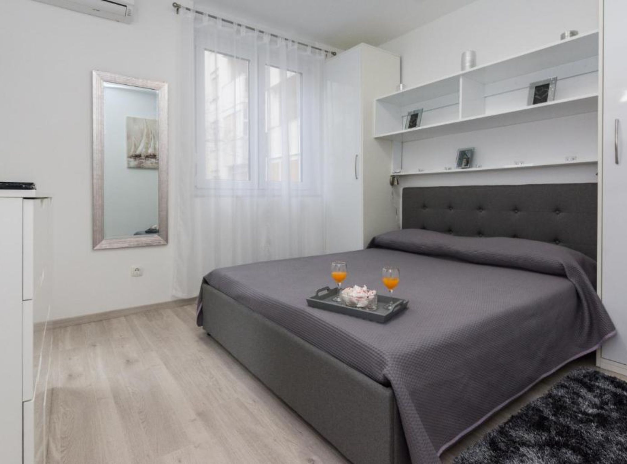 A modern bedroom with a grey bed, a bedside table, a mirror, and a window with sheer curtains.