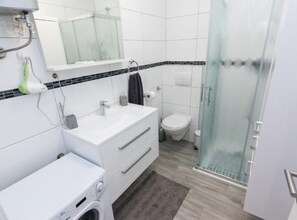 A modern bathroom with a white sink, mirror, and toilet, a glass shower enclosure, and a built-in washing machine. - Stylish Zadar Apartments With Easy Access to Old Town (Zadar)