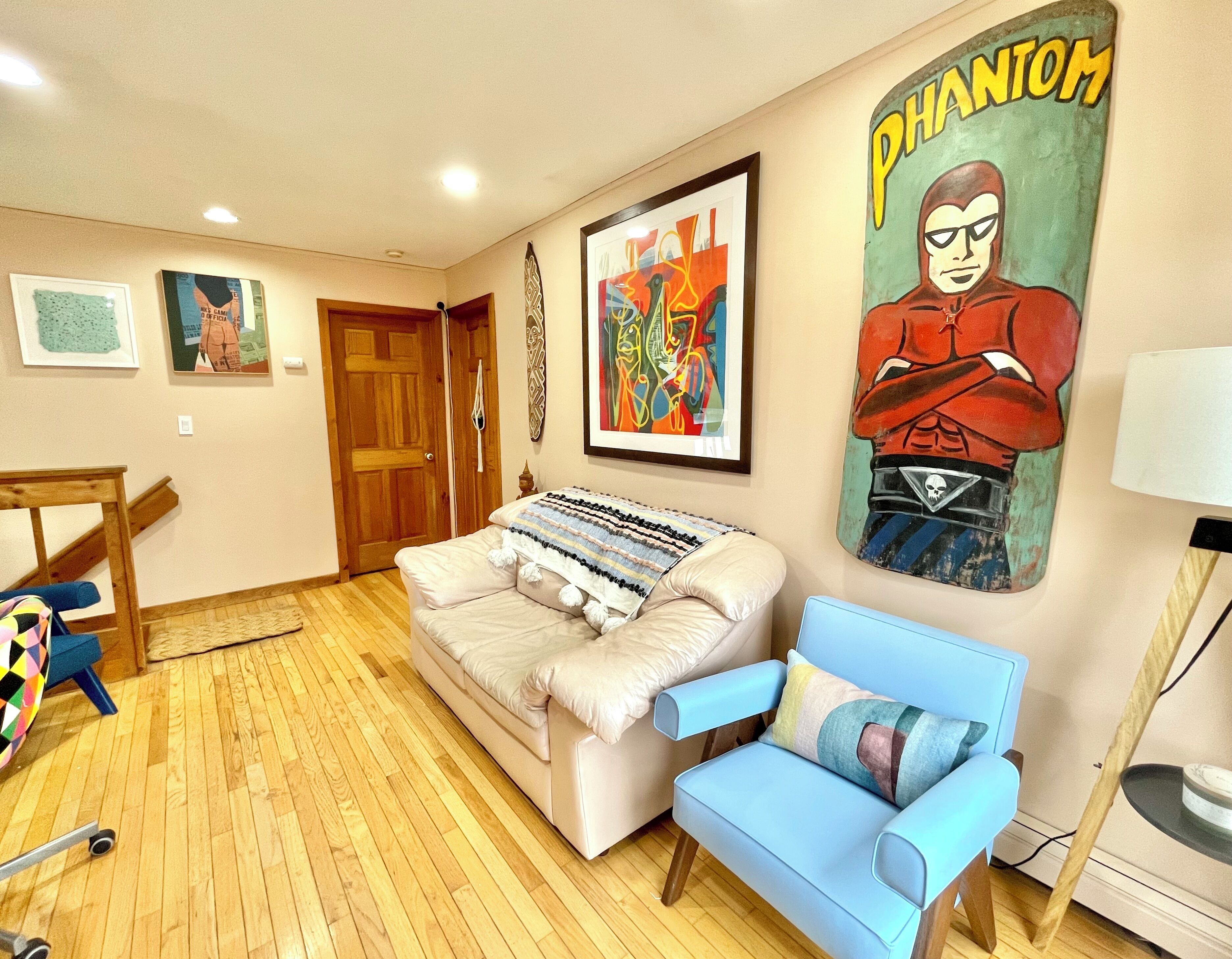 A room with a beige sofa, a blue armchair, a wooden floor, a door, and a wall with a 'Phantom' poster.