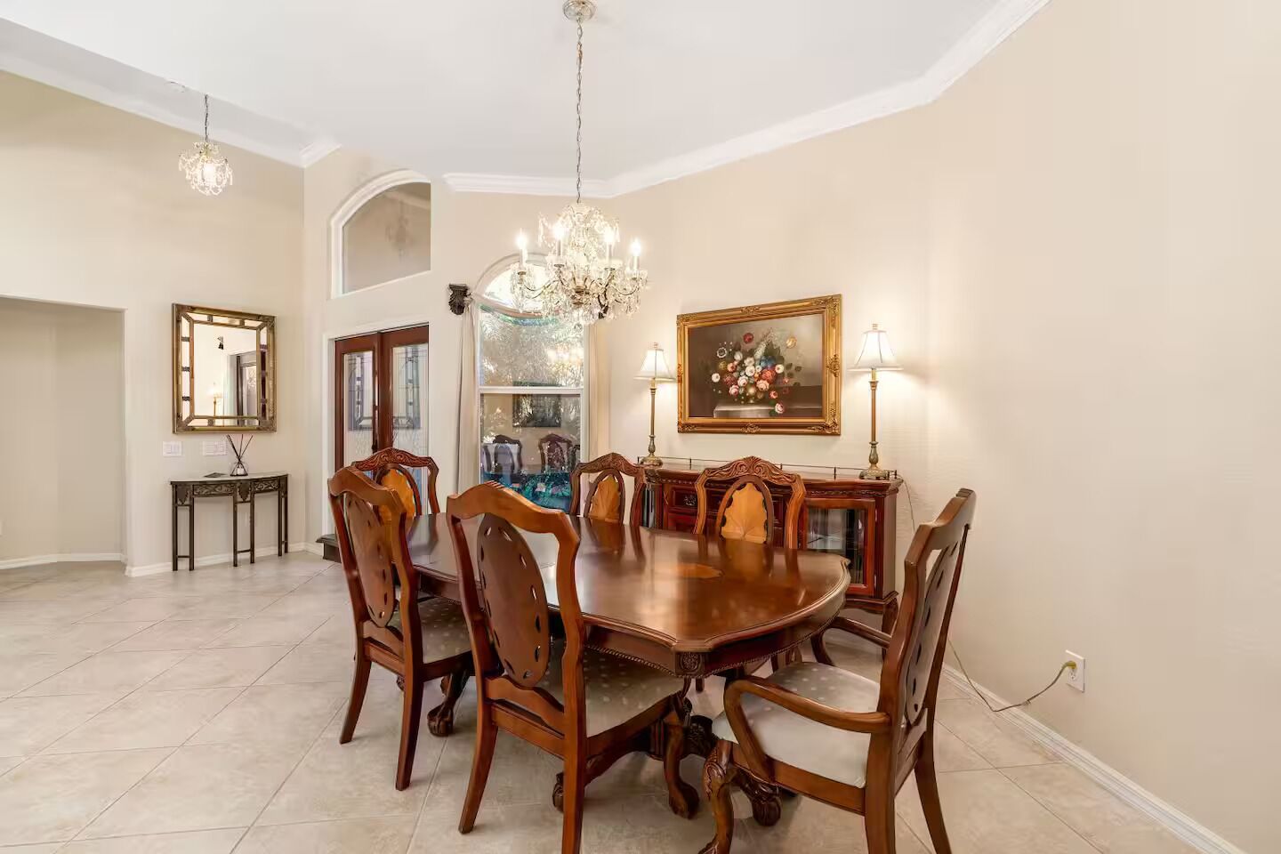 A dining room with a large wooden table, six chairs, a chandelier, a painting, and a console table with a mirror.