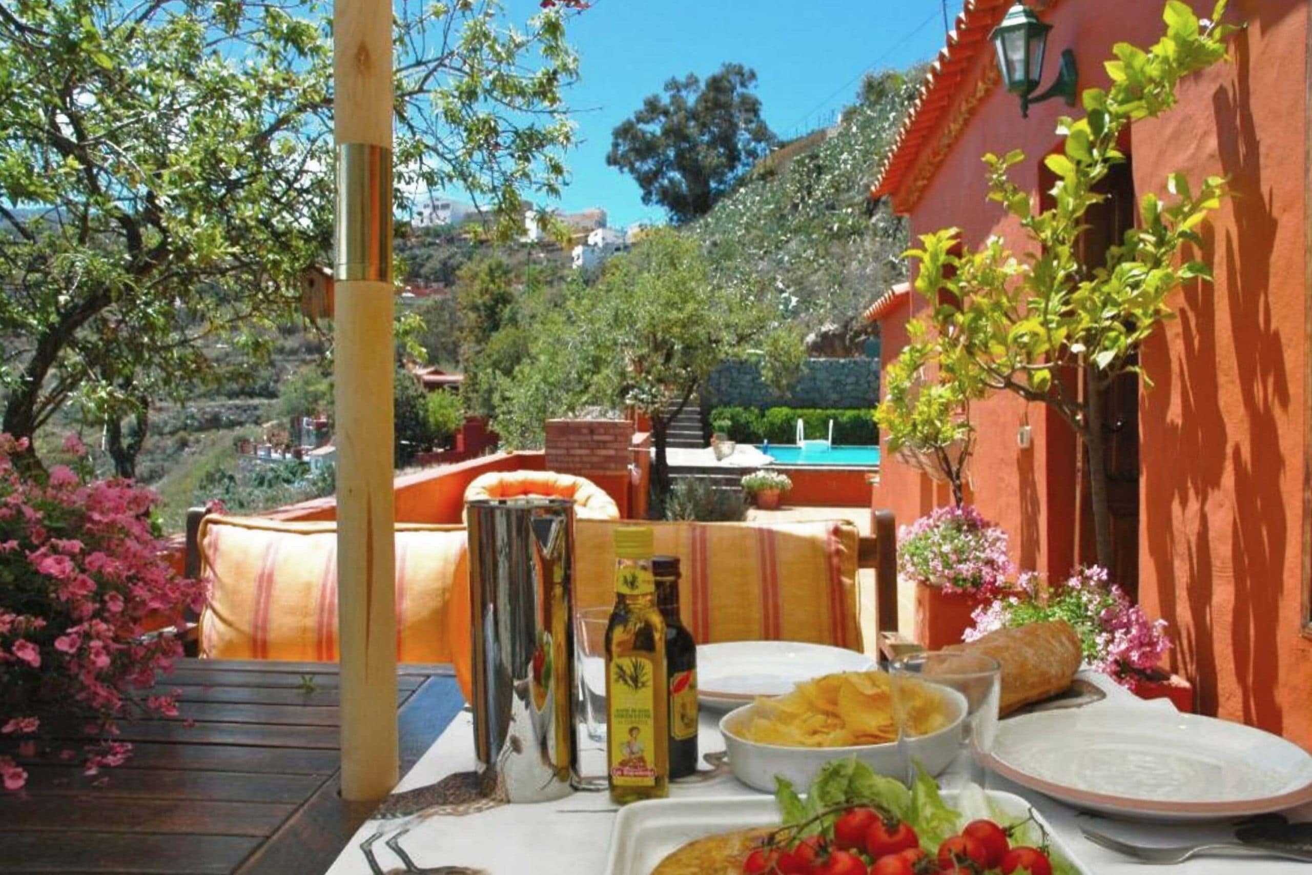 An outdoor dining area with a table set for a meal, surrounded by greenery and a hillside view.