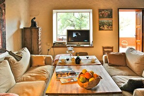 A living room with a sofa, a coffee table with magazines and fruit, a television, and a window with a view of trees. - Casa Tres Almendros (Vega de San Mateo)