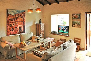 Uma sala de estar acolhedora com um sofá, uma mesa de centro de madeira e uma televisão. Há um quadro na parede e uma janela com vista para o exterior.