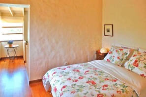 Quarto com colcha florida, uma pequena mesa com uma planta e um quadro na parede.