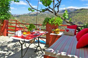 Zona d'estar a l'aire lliure acollidora amb una taula preparada per a una estada, un llit amb almoades vermelles i una vista panoràmica de les muntanyes.
