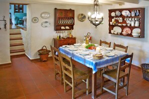Ein Essbereich mit einem für vier Personen gedeckten Tisch, umgeben von Holzstühlen. Eine Kristallleuchter hängt über dem Tisch, und an Wandschränken stehen Geschirr und Teller. Im Hintergrund ist eine Treppe zu einem anderen Raum sichtbar.