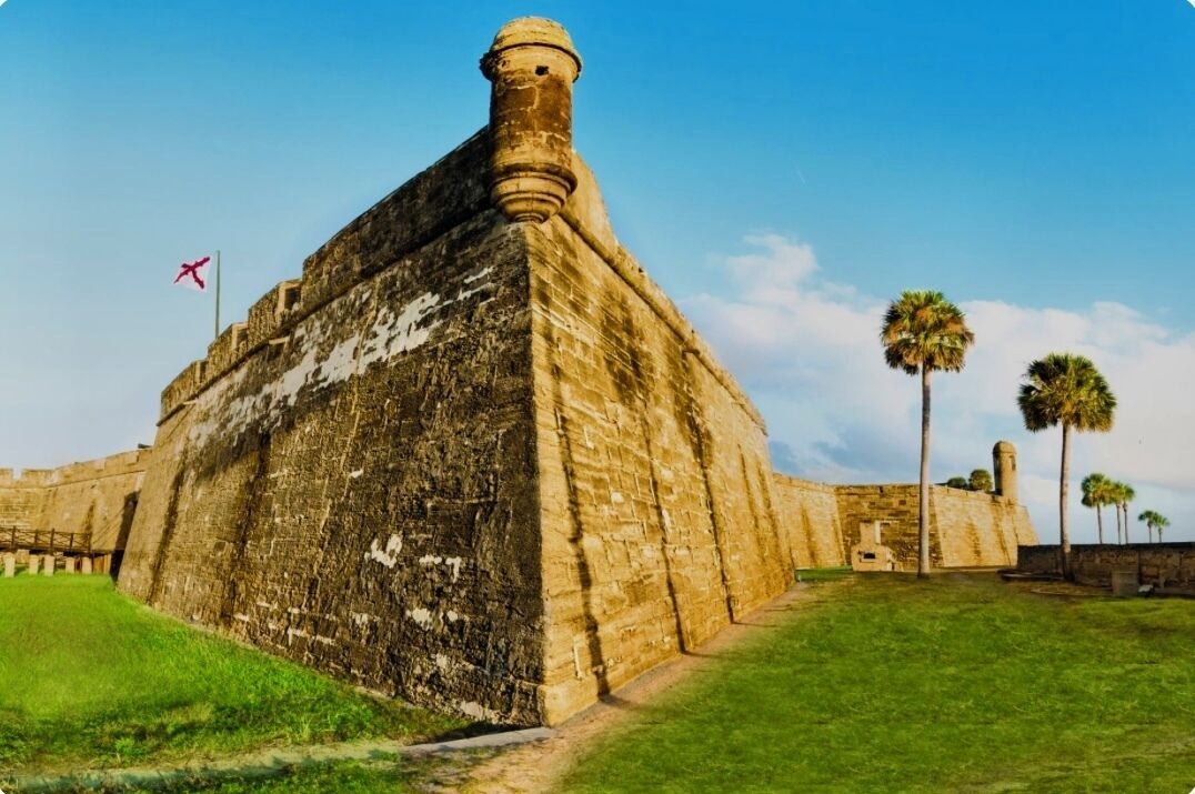 A large, historic stone building with a prominent tower, surrounded by a grassy area and palm trees.