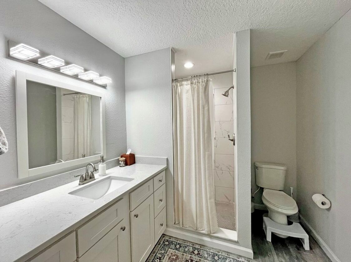 Une salle de bain équipée d’un lavabo, d’un miroir, d’un rideau de douche, de toilettes et d’un petit marchepied.