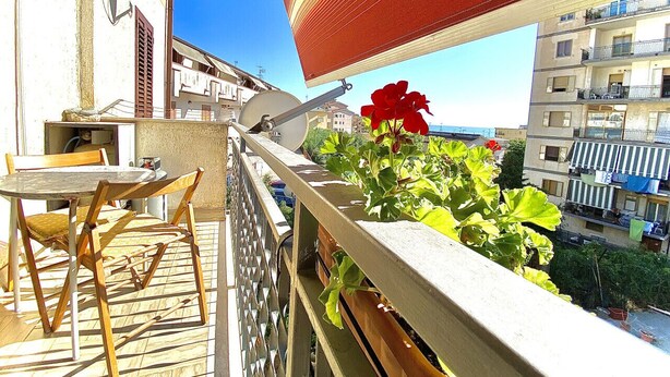 En balkon med et bord og stole, en plantekasse med røde blomster, og udsigt over bybilledet med bygninger.