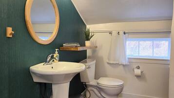 Un baño con lavamanos pedestal, inodoro, repisa de madera con una planta y ventana con cortina.