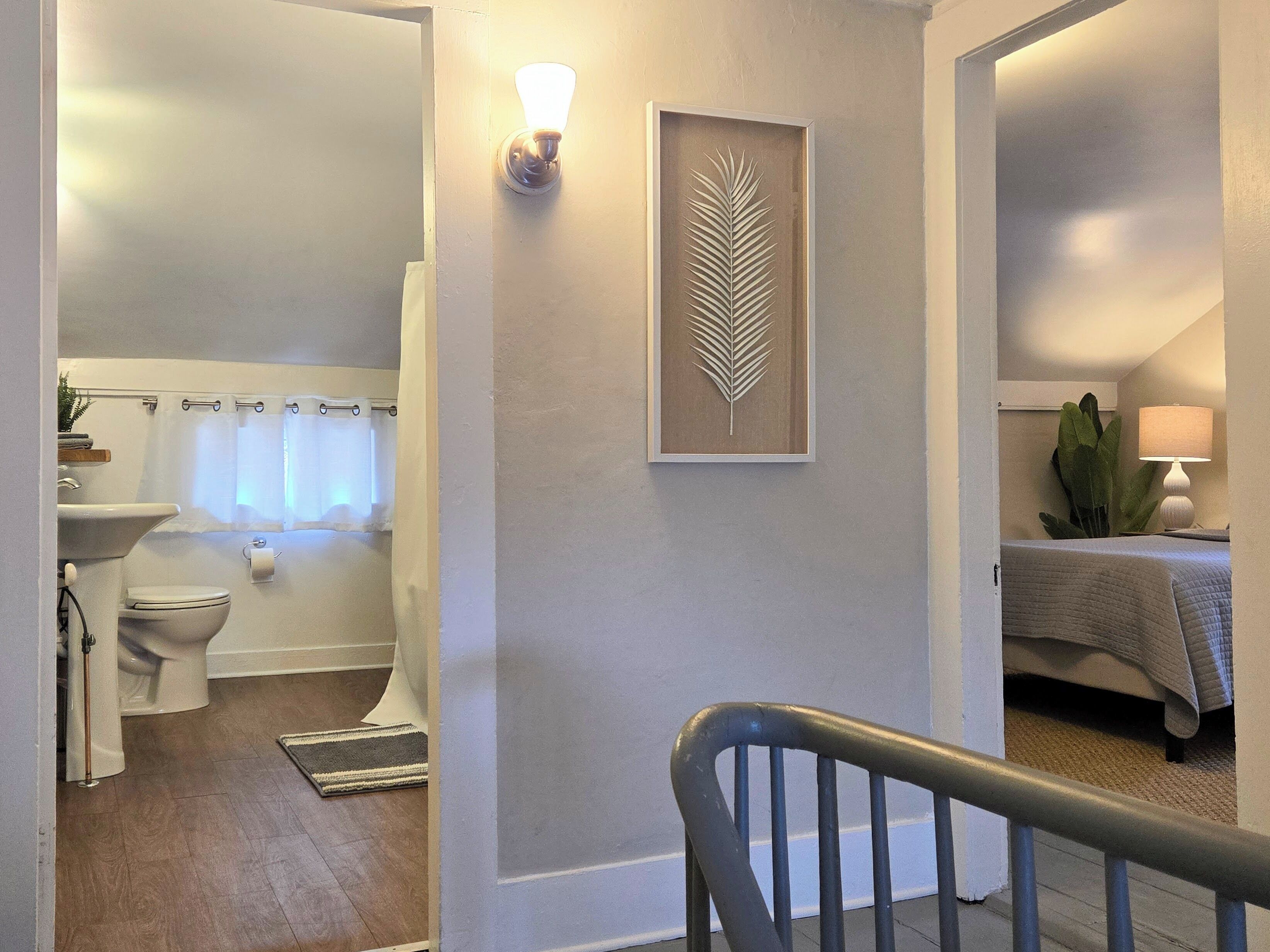 A bathroom with a toilet, sink, and shower curtain. There is a framed leaf decoration on the wall and a lamp on a bedside table.