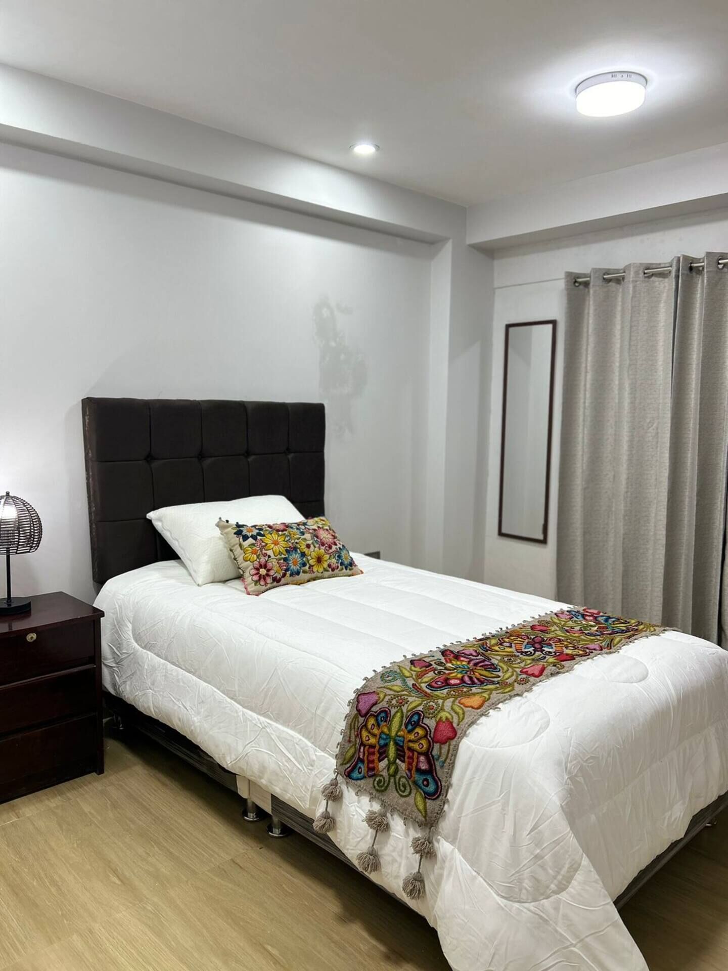 A neatly made bed with a white duvet and colorful pillows, a dark headboard, a bedside lamp, and a curtain on the window.